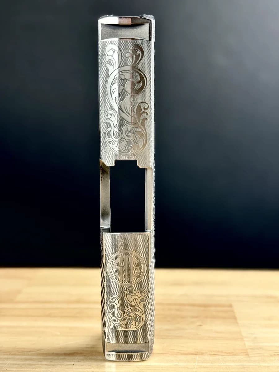 Engraved pistol slide with scrollwork and gold inlay, held in a vise